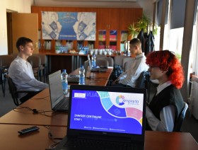 troje zawodników w świetlicy urzędu, na pierwszym planie monitor, na którym wyświetla się grafika promująca olimpiadę